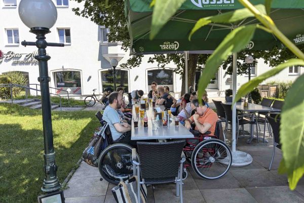 eine Gruppe Erwachsener mit Behinderungen sitzen an einem Tisch in einem Gastgarten; es stehen Getränke und Speisen am Tisch