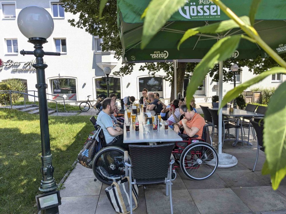 eine Gruppe Erwachsener mit Behinderungen sitzen an einem Tisch in einem Gastgarten; es stehen Getränke und Speisen am Tisch